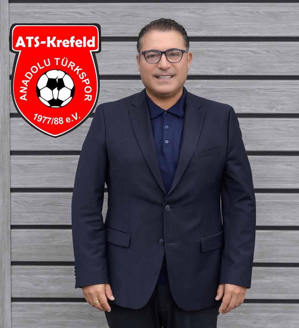 Mann in Anzug mit Brille steht vor grauer Wand, links daneben Vereinslogo von Anadolu T&uuml;rkspor.