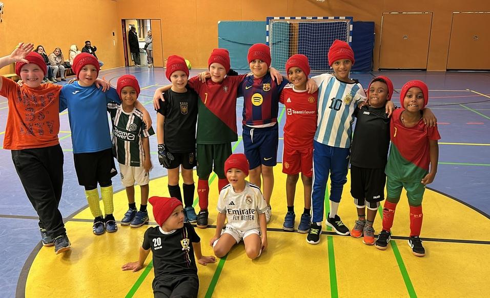 Gruppe von Kindern in Sportkleidung mit roten M&uuml;tzen, posierend auf einem bunten Sportboden in einer Halle.