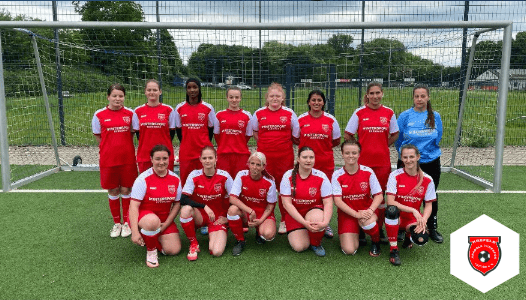 Mannschaftsfoto einer Frauenfu&szlig;ballmannschaft in roten Trikots vor einem Tor auf einem Kunstrasenplatz.