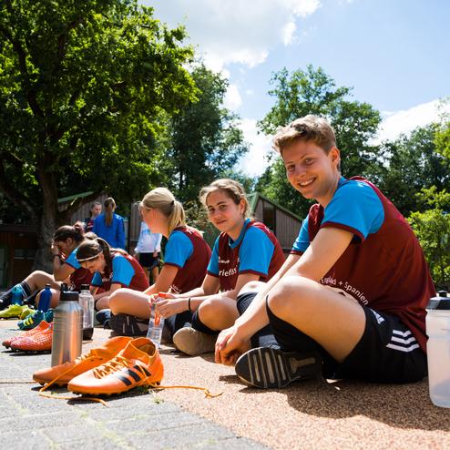 Gruppe junger Fu&szlig;ballspielerinnen sitzt auf dem Boden, w&auml;hrend sie ihre Schuhe schn&uuml;ren, umgeben von B&auml;umen und Sonne.