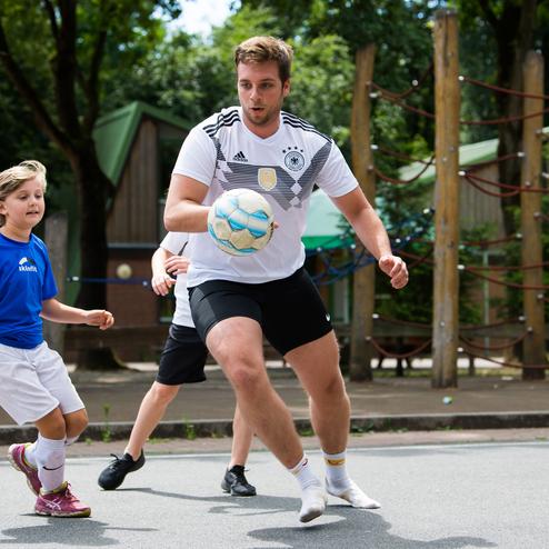 Ein junger Mann dribbelt einen Fu&szlig;ball, w&auml;hrend ein M&auml;dchen in blauem Trikot ihm folgt. Spielszene im Freien.