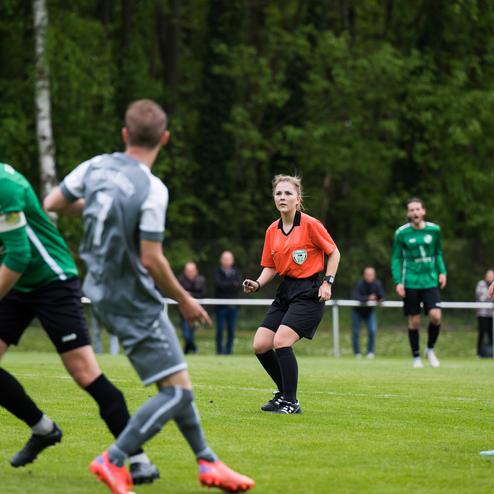 Fu&szlig;ballspiel mit Spielern in grauen und gr&uuml;nen Trikots sowie einer Schiedsrichterin in Orange auf einem Rasenplatz.