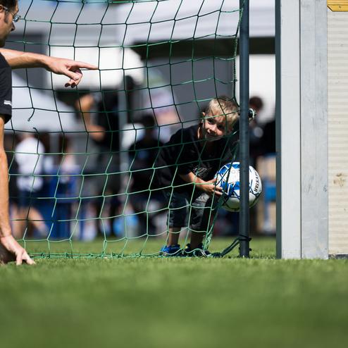 Ein Kind schl&uuml;pft durch ein Fu&szlig;balltor und h&auml;lt einen Ball, w&auml;hrend ein Erwachsener es ermutigt.
