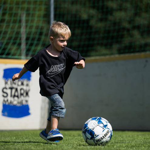 Ein kleiner Junge in einem schwarzen T-Shirt spielt mit einem Fu&szlig;ball auf einem gr&uuml;nen Rasenfeld.
