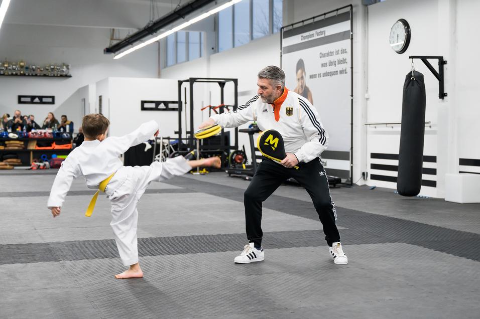 Junge in Karateanzug mit gelbem Gürtel führt einen Kick gegen gepolsterte Schlaghandschuhe eines Trainers aus.